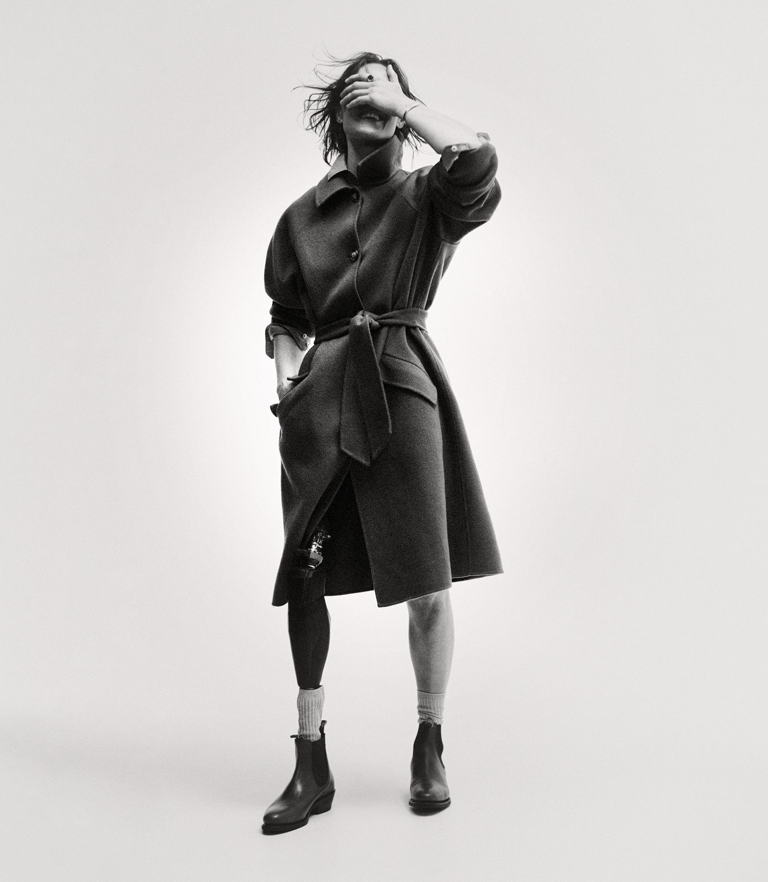 Monochrome portrait of a person wearing an overcoat with a belt and boots, shielding their eyes with one hand in a studio setting.