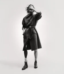 Model wearing a dark belted coat and boots with one hand covering eyes against a plain white background