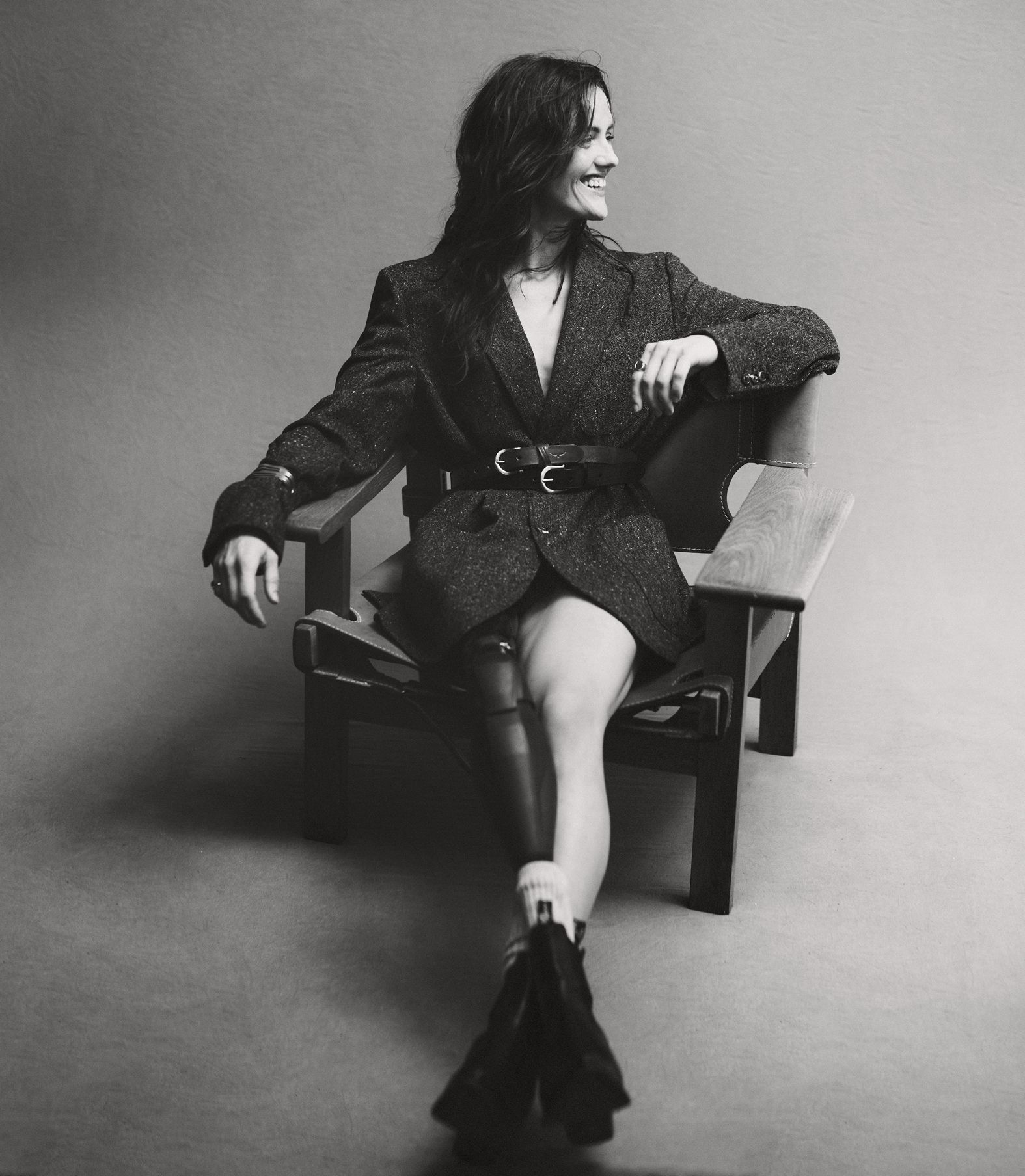 Monochromatic studio portrait of a woman sitting in a chair, wearing a tweed jacket, smiling and looking away.