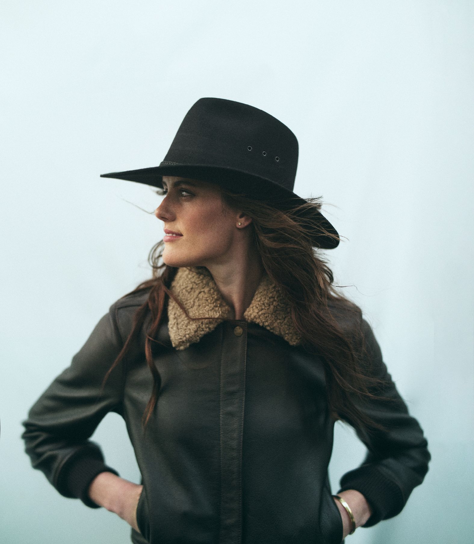 Stylish woman in a black hat and brown leather jacket poses against a light background in a medium shot, looking off to the side.