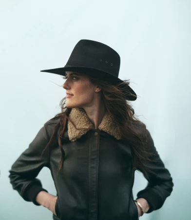 Stylish woman in a black hat and brown leather jacket poses against a light background in a medium shot, looking off to the side.