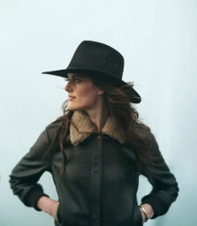 Woman wearing a black fedora hat and a brown leather jacket with shearling collar against a light background