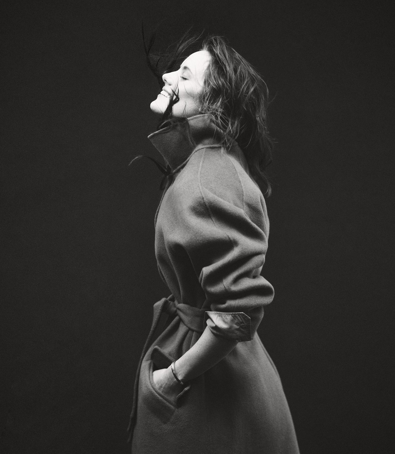 Monochrome portrait of a smiling woman in a trench coat, eyes closed, wind blowing her hair against a dark, neutral background.