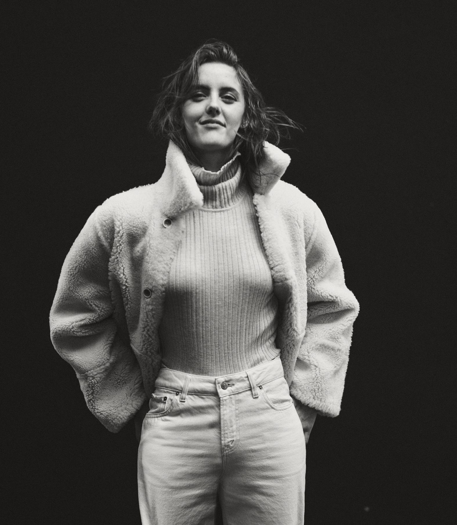Portrait of a young woman with wavy dark hair, wearing a fuzzy coat and jeans, against a dark backdrop in soft, diffused light.