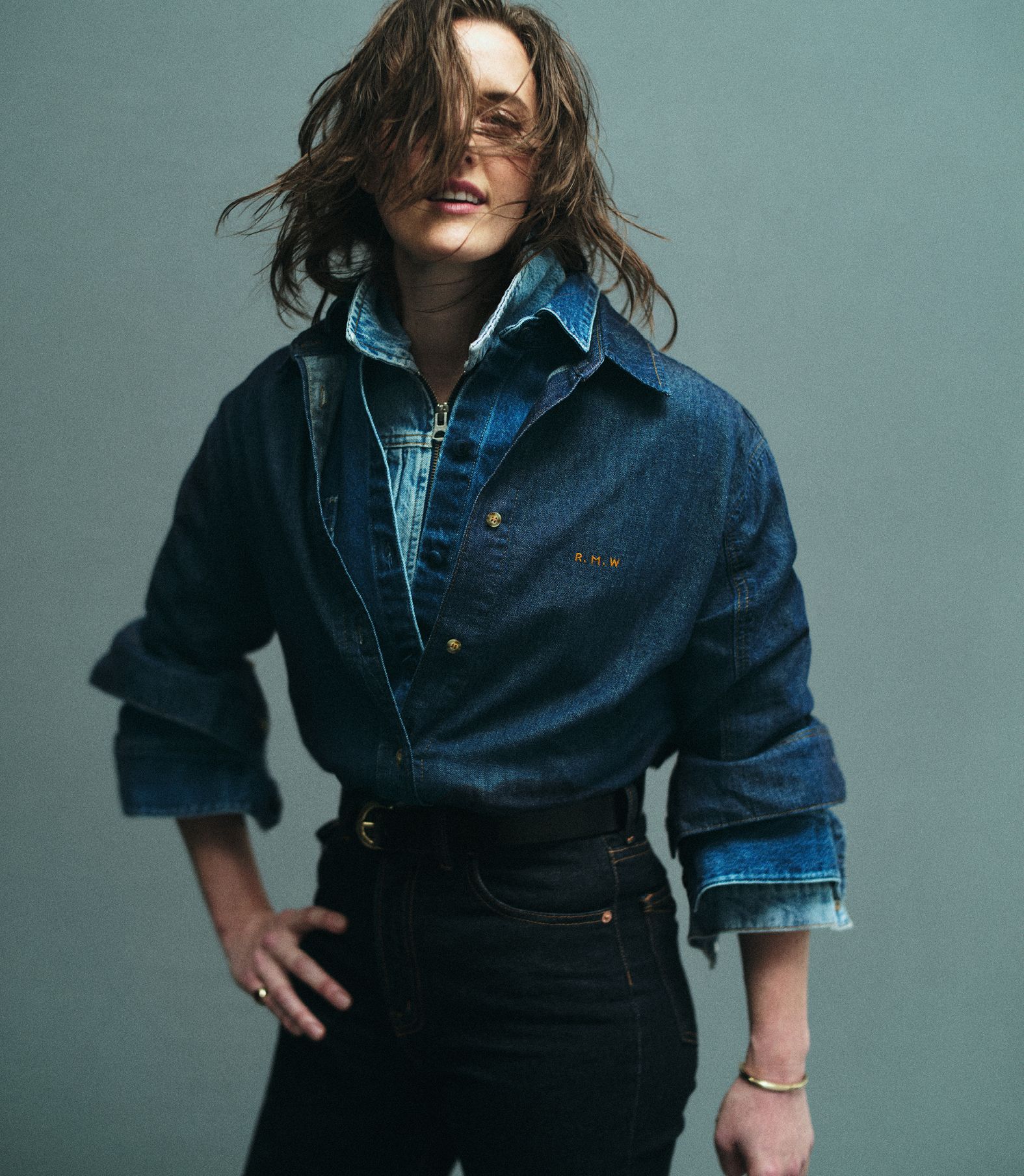 A woman poses indoors wearing a denim top and black jeans, standing against a grey background in a studio shot. 