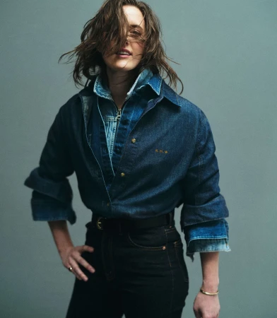 A woman poses indoors wearing a denim top and black jeans, standing against a grey background in a studio shot. 
