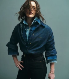 Woman wearing layered denim shirts and dark jeans posing against a plain grey studio backdrop