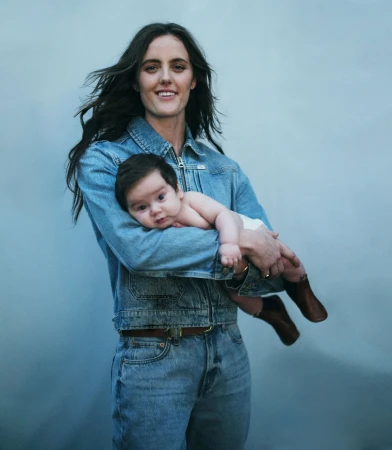 A woman in a denim jacket holds a baby in a diaper, both smiling, against a muted blue background in a portrait.