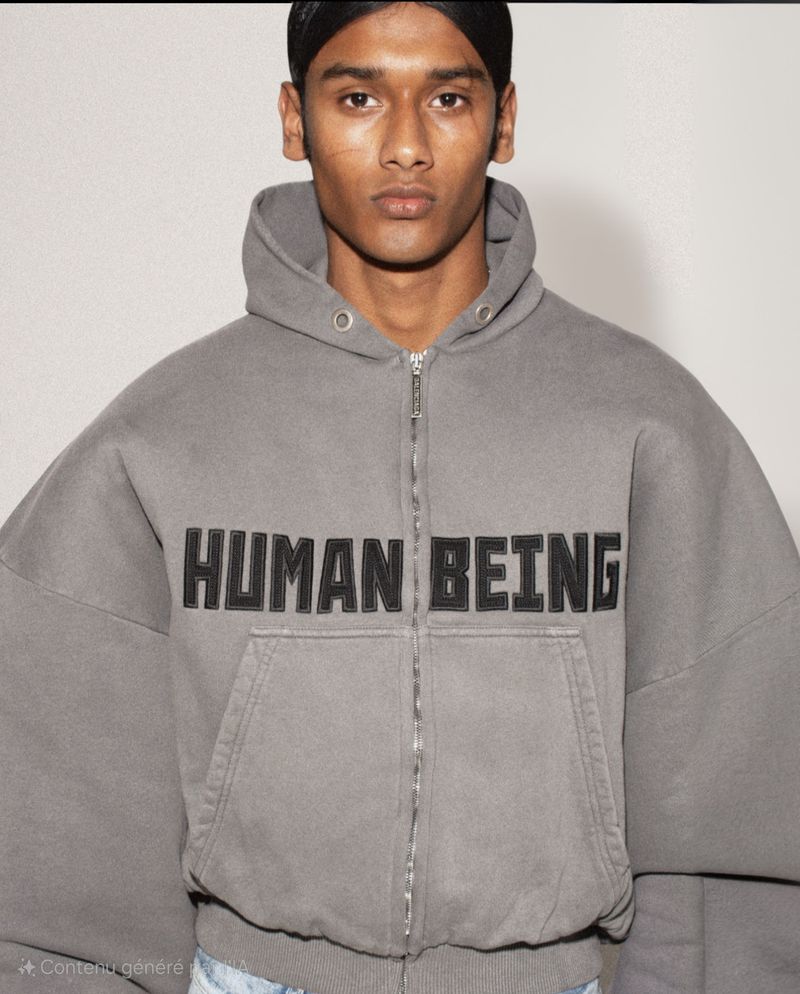Close-up of an adult male wearing a gray, zip-up hoodie with 'HUMAN BEING' on the front, set against a neutral backdrop.