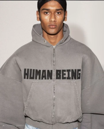 Close-up of an adult male wearing a gray, zip-up hoodie with 'HUMAN BEING' on the front, set against a neutral backdrop.