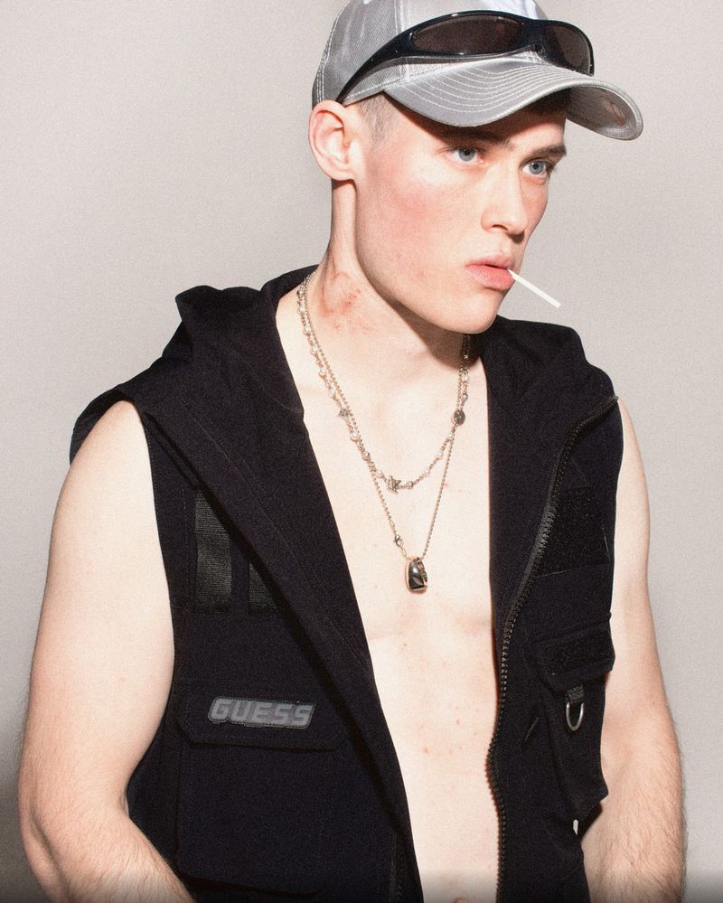 Portrait of a male adult wearing a black vest and baseball cap with sunglasses, sucking on a lollipop in a studio setting.