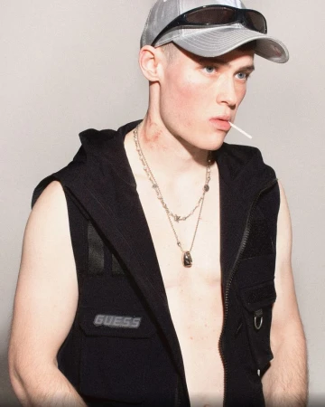 Portrait of a male adult wearing a black vest and baseball cap with sunglasses, sucking on a lollipop in a studio setting.