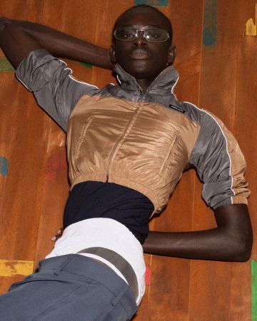A Black male model poses indoors wearing sunglasses, a cropped brown and gray jacket, a black shirt, and blue pants against a textured backdrop.