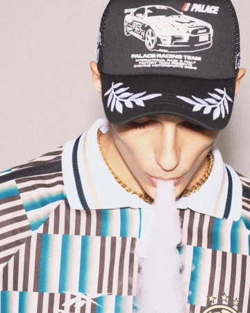 Young man in Palace Racing Team cap exhales vapor, striped shirt, and gold chain against a neutral backdrop, captured in a studio setting.