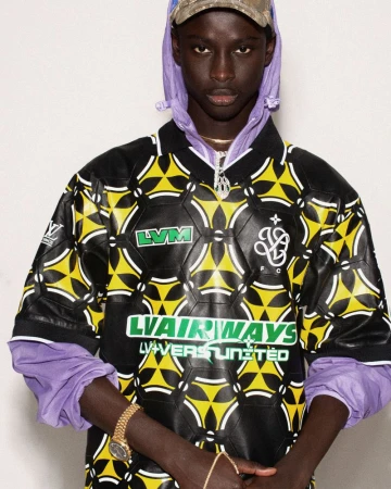 Male fashion model poses in a black and yellow patterned collared shirt with logo detail, light purple hoodie, and camouflage cap indoors.