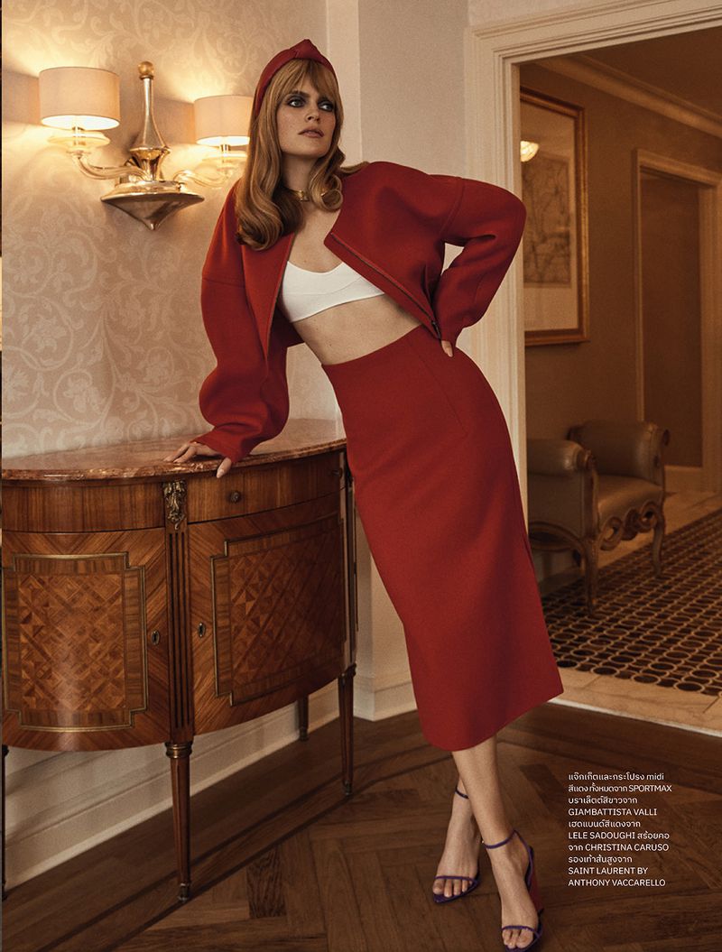 A stylish adult woman poses indoors wearing a red skirt and jacket with a white top and violet heels against a vintage cabinet and wall.