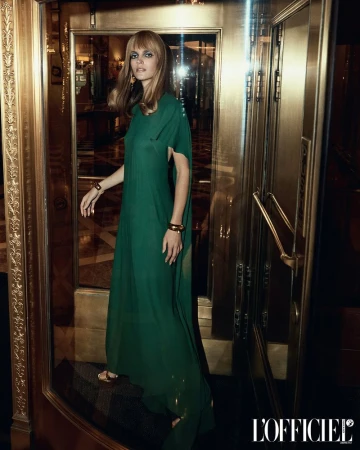 Fashion photo of a blonde model in a long green dress, gold bracelets, and gold earrings, standing in a gold-toned doorway.