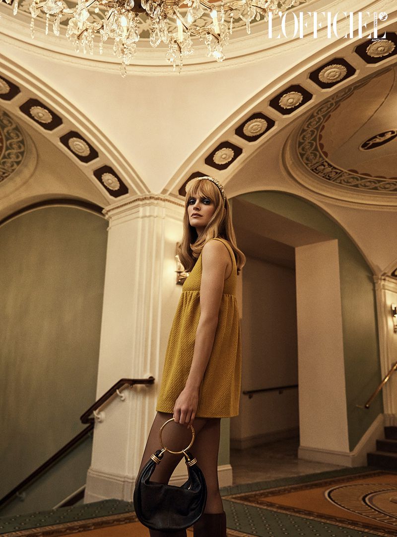 A stylish adult woman wearing a yellow 60s-inspired mini dress, tights, and boots poses with a handbag on a staircase indoors.