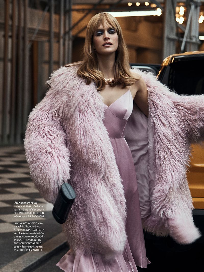 Fashion shot of a woman in a pink dress and faux fur coat standing on a city street with a yellow taxi in the background.
