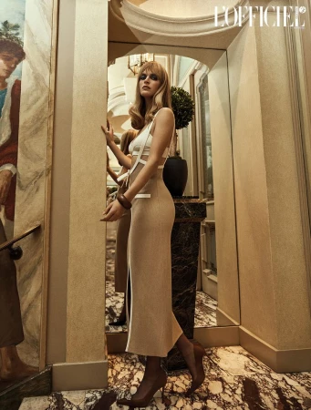 A blonde woman in a cut-out white top and beige dress poses indoors next to a mirror and a painted wall, showcasing fashion and style.