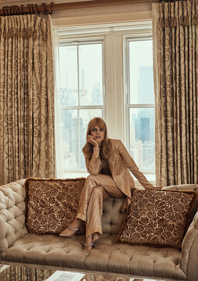 A woman in a shimmering suit sits on a tufted sofa near a window with city views, bathed in soft light from a high angle.