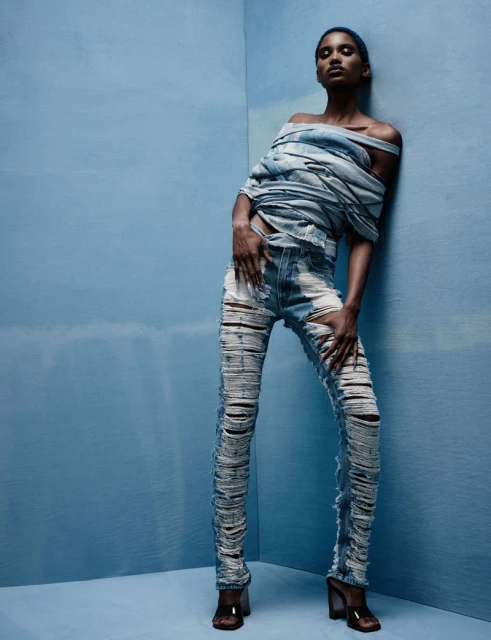 Fashion shot of a slender Black woman in ripped denim jeans and a deconstructed denim top, posing in a light-blue corner studio.