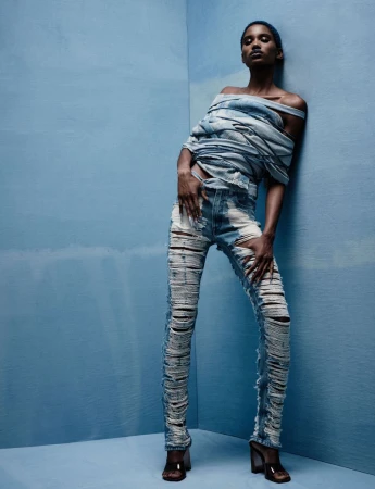Fashion shot of a slender Black woman in ripped denim jeans and a deconstructed denim top, posing in a light-blue corner studio.
