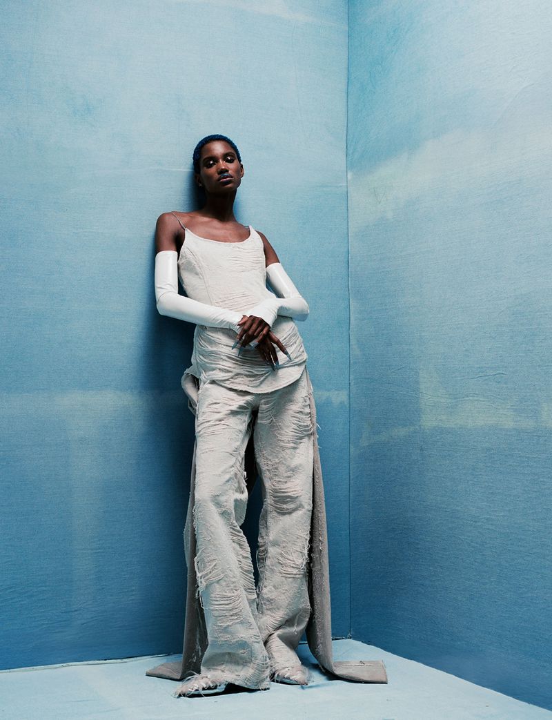 A slender African-American woman in a light beige tank top and distressed pants with train stands indoors against a mottled blue wall.