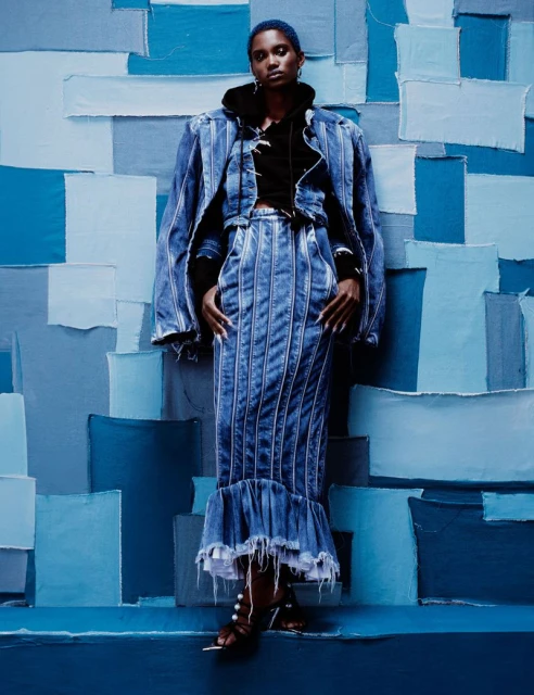 Fashion editorial shot of a black female model in a full denim outfit against a textured denim backdrop in a studio setting.