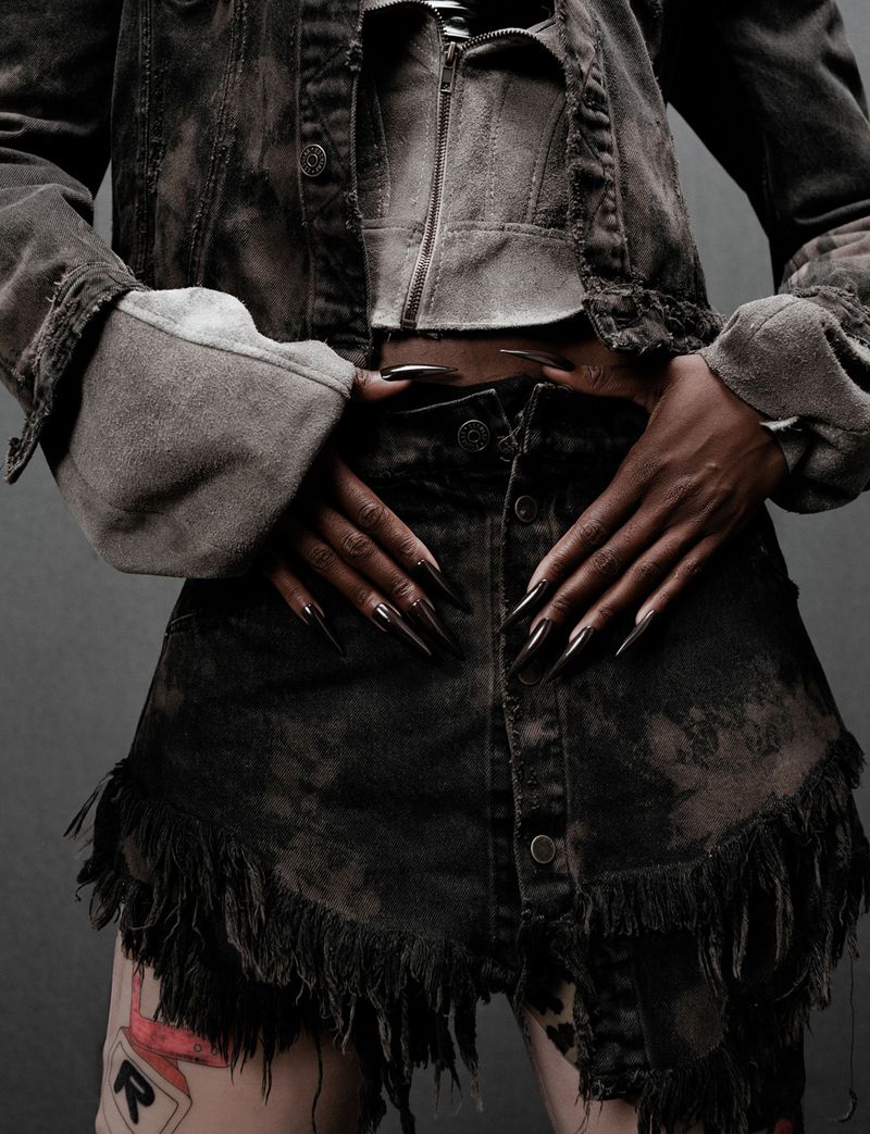 Woman poses in distressed denim jacket and skirt with fringe detail, showcasing long black stiletto nails against a grey backdrop.