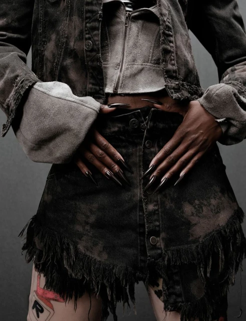 Woman poses in distressed denim jacket and skirt with fringe detail, showcasing long black stiletto nails against a grey backdrop.