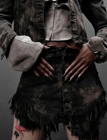 Woman poses in distressed denim jacket and skirt with fringe detail, showcasing long black stiletto nails against a grey backdrop.
