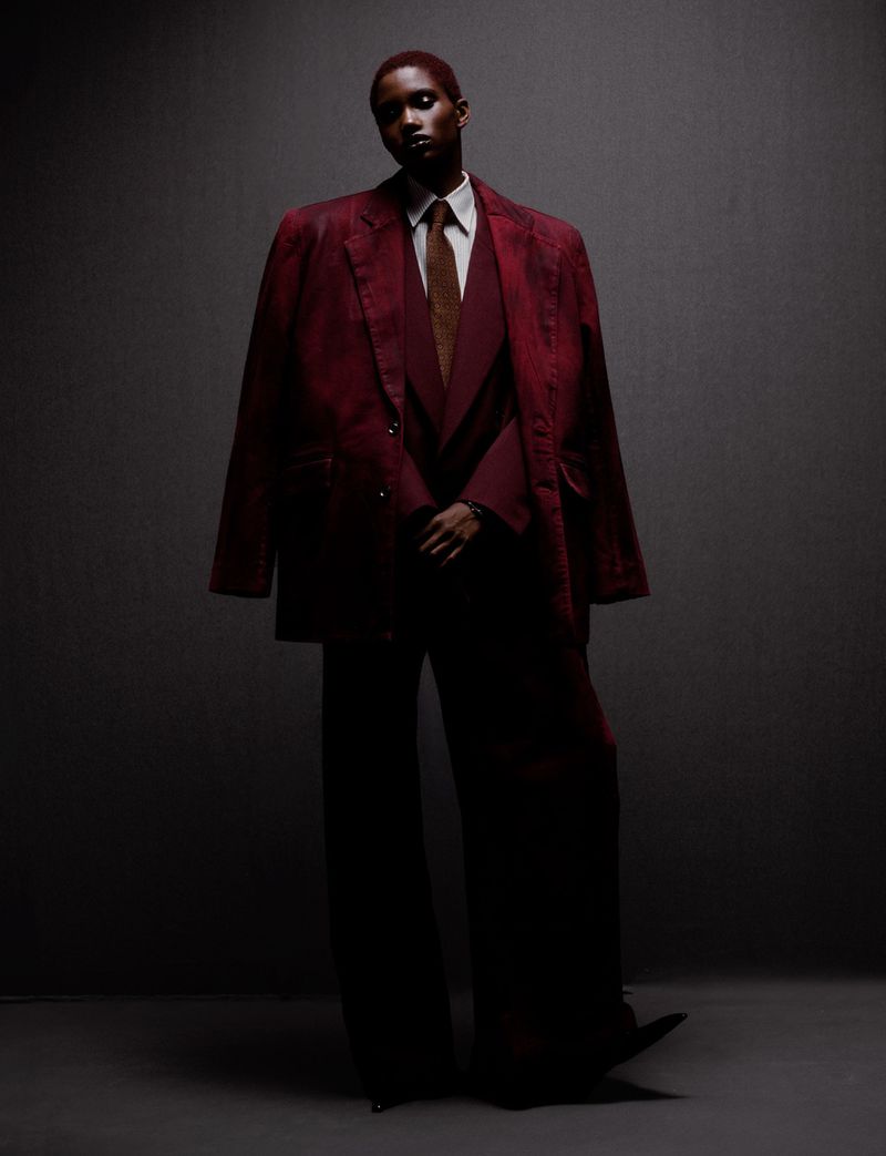 Elegant androgynous fashion portrait of a person with dark skin and red hair wearing a red suit and tie in a dimly lit studio.