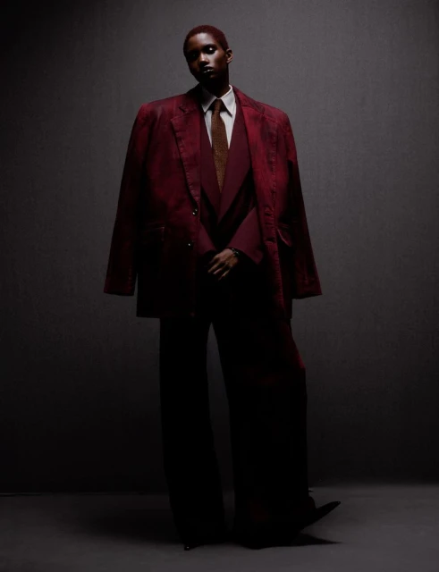 Elegant androgynous fashion portrait of a person with dark skin and red hair wearing a red suit and tie in a dimly lit studio.