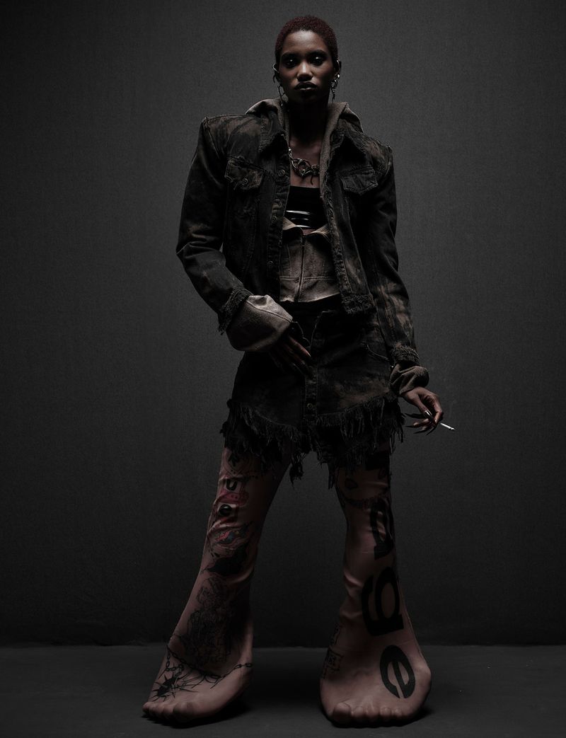 A slender black woman in distressed denim jacket, skirt, and printed tights strikes a confident pose in a dimly lit studio setting.