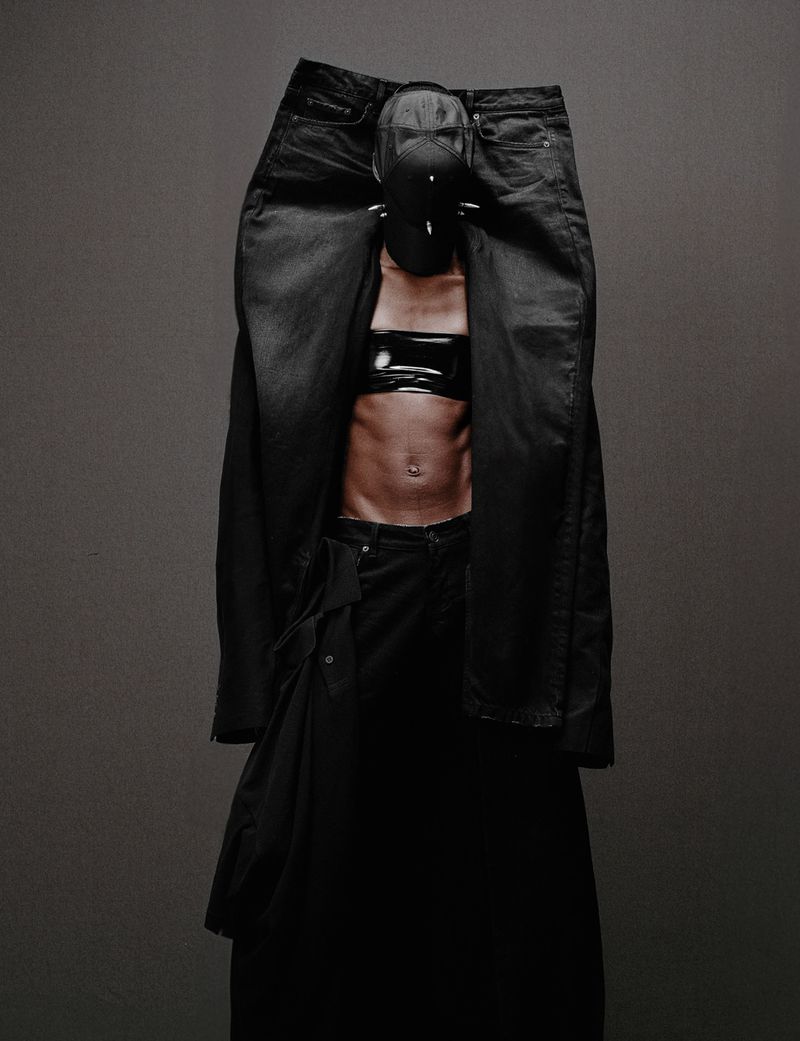 Abstract fashion portrait with an adult wearing black denim, a cropped top, and a spiked mask, posing in front of a grey backdrop.