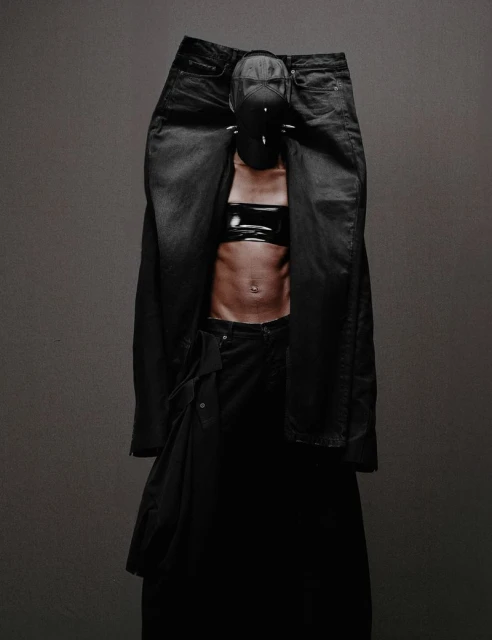 Abstract fashion portrait with an adult wearing black denim, a cropped top, and a spiked mask, posing in front of a grey backdrop.