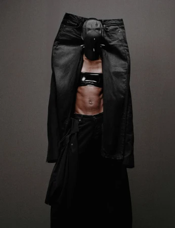 Abstract fashion portrait with an adult wearing black denim, a cropped top, and a spiked mask, posing in front of a grey backdrop.