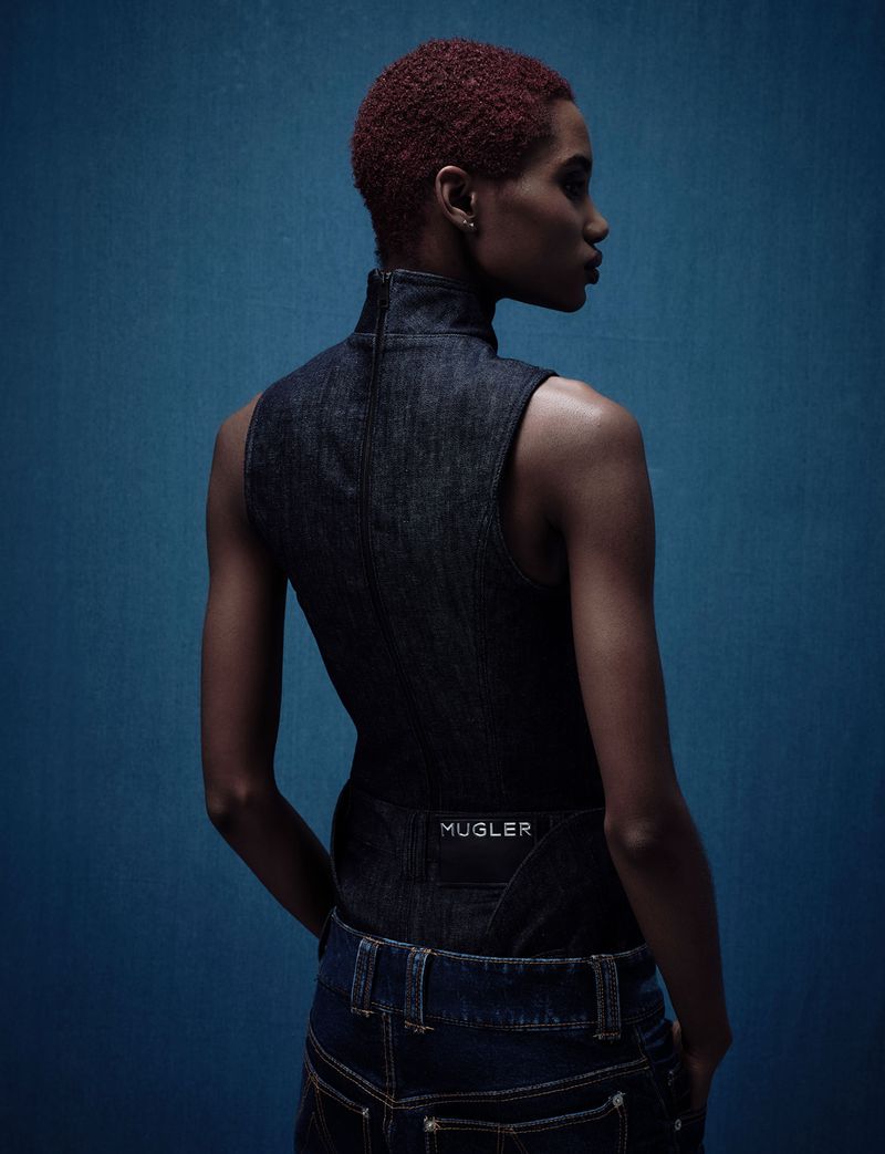 A woman with red hair poses in a dark denim top and jeans against a blue background in a fashion editorial image.