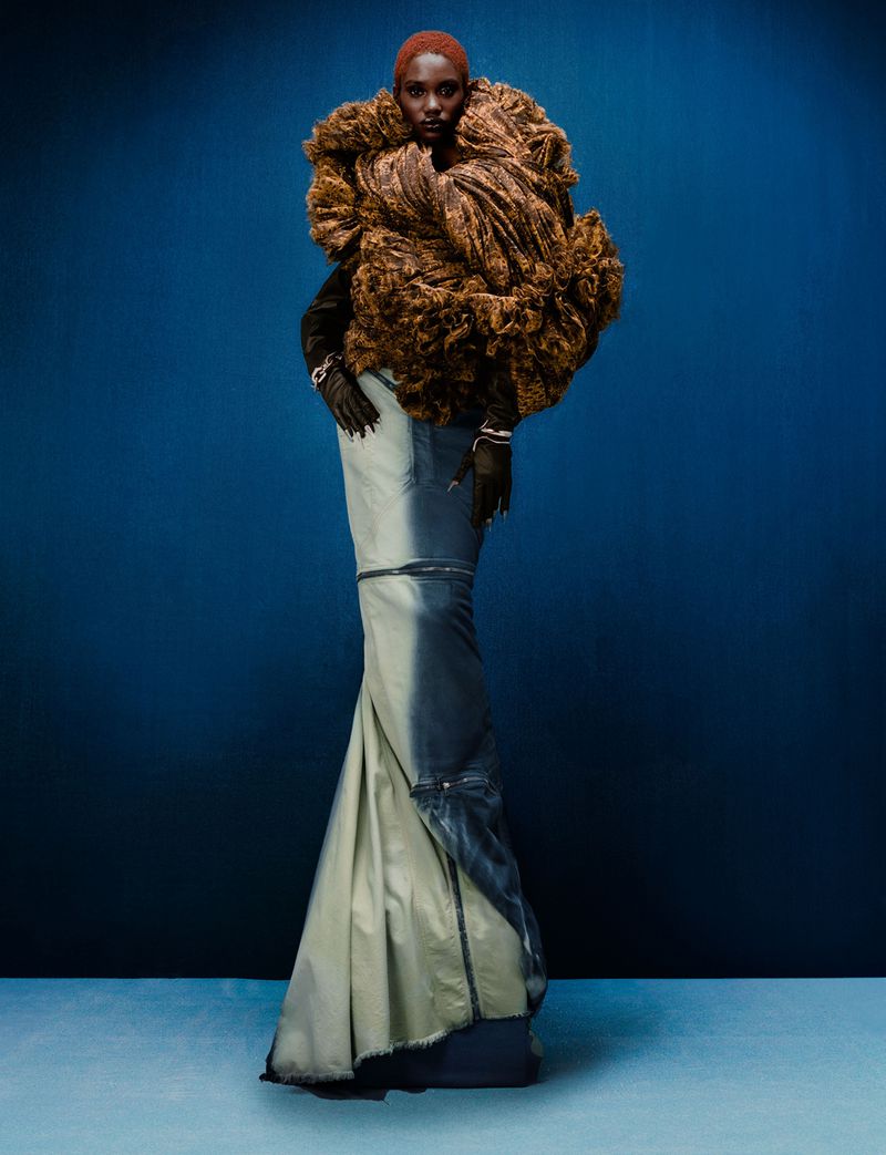 Fashion model in a couture dress with a brown textured collar, posing in front of a solid blue backdrop for a studio fashion editorial.