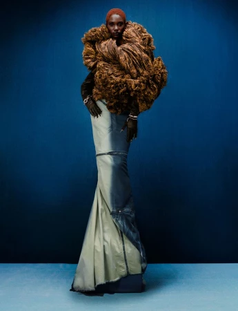 Fashion model in a couture dress with a brown textured collar, posing in front of a solid blue backdrop for a studio fashion editorial.