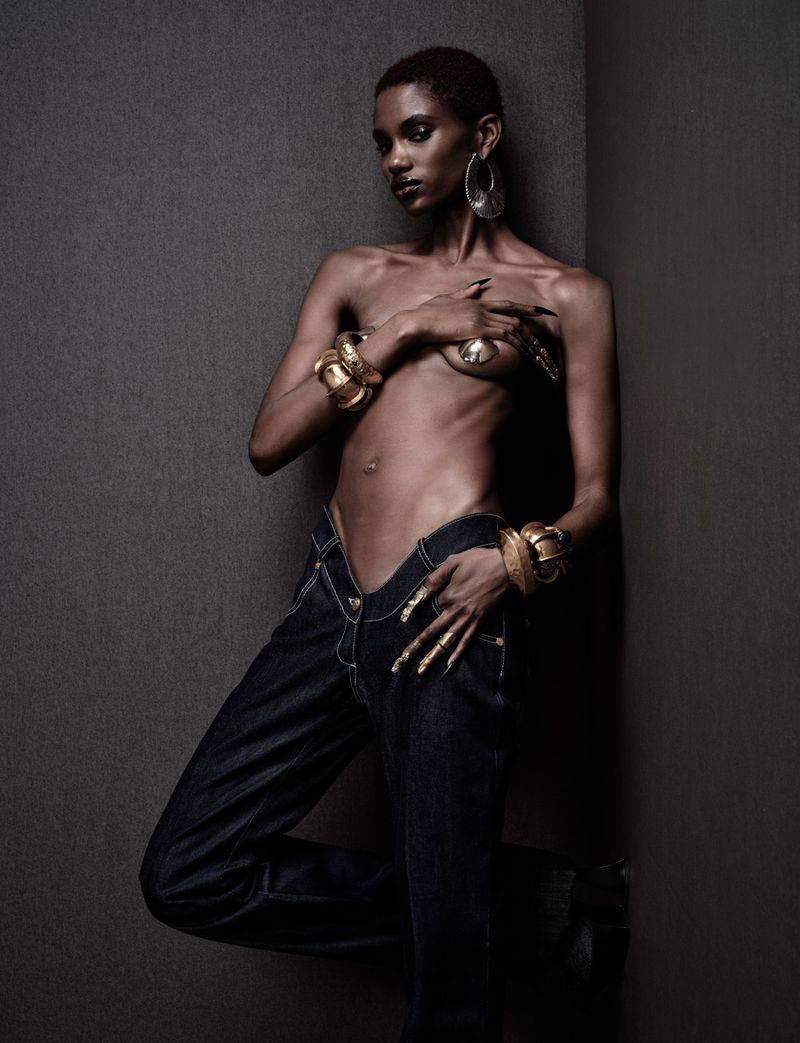 A bare-chested, tan-skinned fashion model with gold jewelry poses against a gray wall wearing dark-wash jeans in a fashion shot.