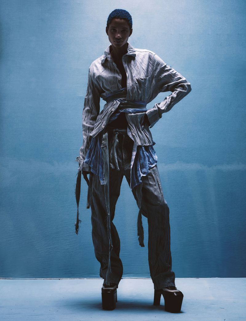 Woman in deconstructed light blue and grey striped shirt and pants, with platform heels, poses against a blue backdrop in a studio.