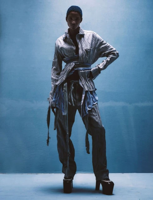 Woman in deconstructed light blue and grey striped shirt and pants, with platform heels, poses against a blue backdrop in a studio.