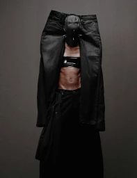 Model wearing a black cap and holding up large black denim jeans against a solid dark grey background.