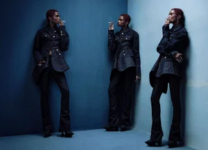 Model with red hair wearing dark denim jacket and pants standing in blue corner holding cigarette