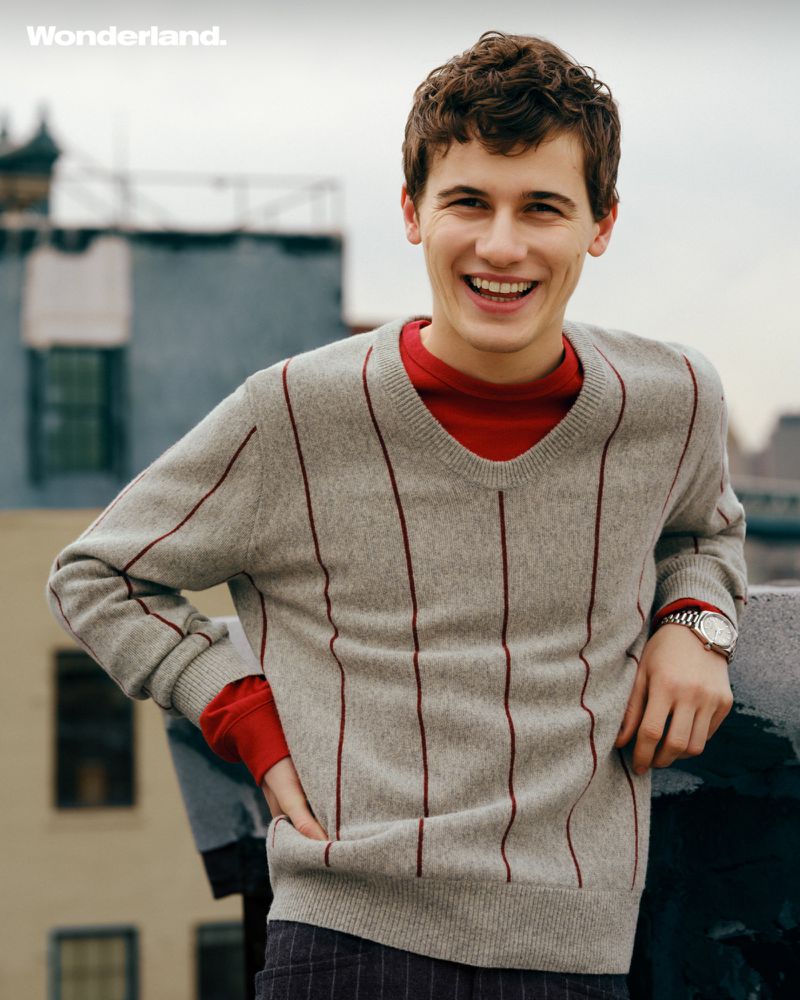 Portrait of actor Sam Nivola smiling, wearing a striped grey sweater over a red top on a rooftop for Wonderland Magazine.