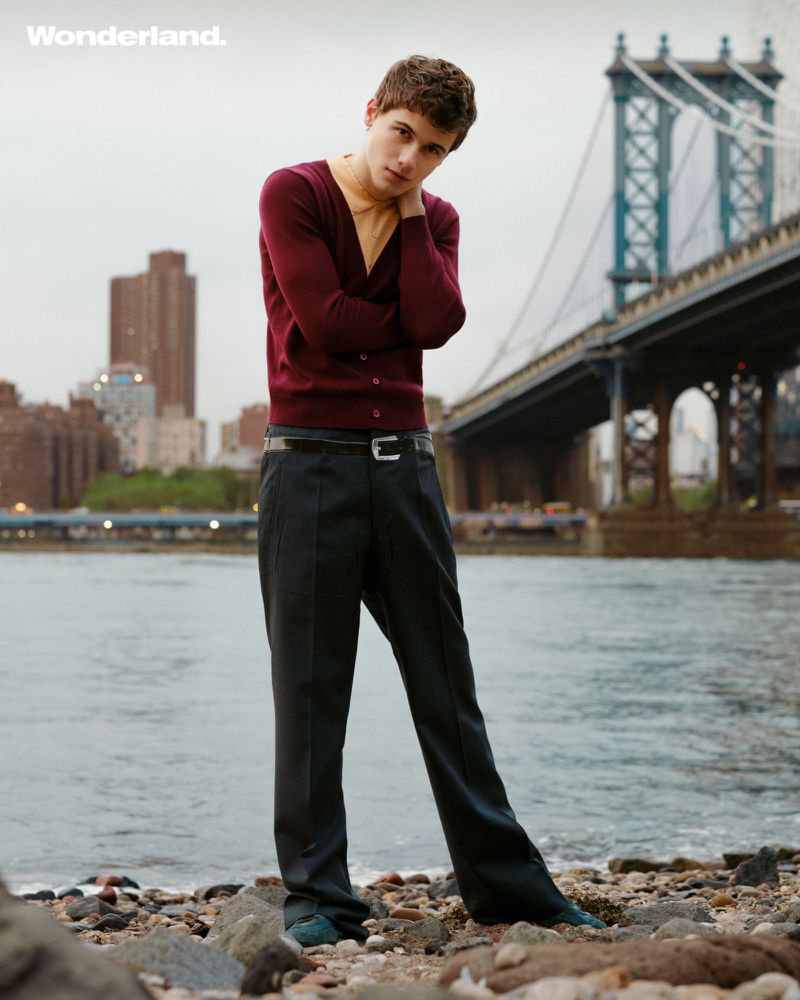 A young adult male poses near the water with a bridge and city skyline in the background, wearing a cardigan and slacks.