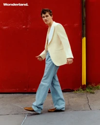 Sam Nivola wearing a cream blazer and blue jeans walking past a bright red wall for Wonderland Magazine editorial.