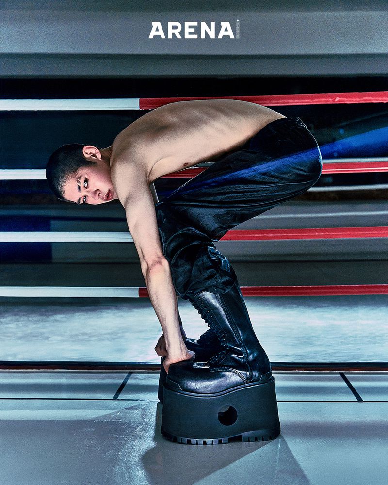 Shirtless person in black pants and platform boots bends over on elevated platform in indoor space with 'Arena' logo sign.